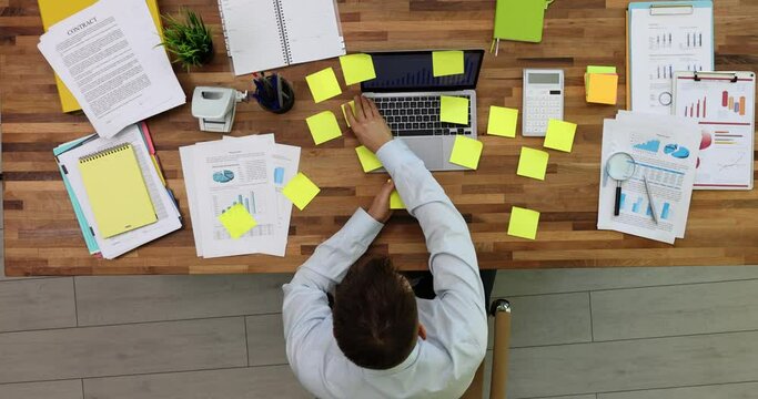 Tired exhausted businessman working on laptop in office with many inscriptions on stickers, top view. Freelancer man busy with work feels multitasking and deadline