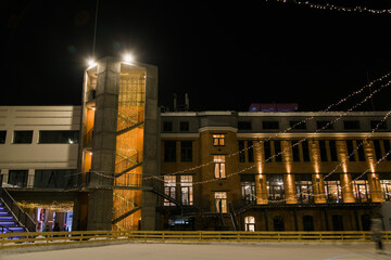 Facade of brick building with outdoor iron stairs with lights and illuminations. Industrial building converted into offices and retail space. And in front of that is a skating rink