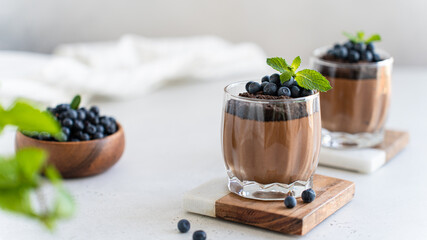 Vegan chocolate mousse in glasses decorated with fresh blueberry and mint on a marble board on a white concrete background. Close up. Protein diet recipe. Creamy chocolate yogurt, panna cotta dessert - obrazy, fototapety, plakaty