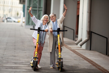 Portrait happy smiling gray haired Senior women enjoying ride together on electric scooters outdoors. Two retired female standing on city street and wave hands