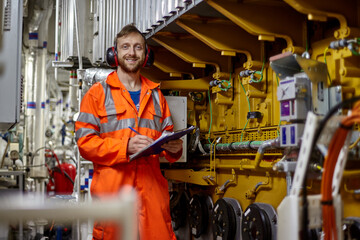 Portrait of young engineer with smile in orange coverall  doing check list in engine room. - obrazy, fototapety, plakaty
