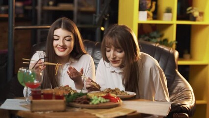 Two beautiful girls in a restaurant at a table are smiling and eating Thai food with chopsticks