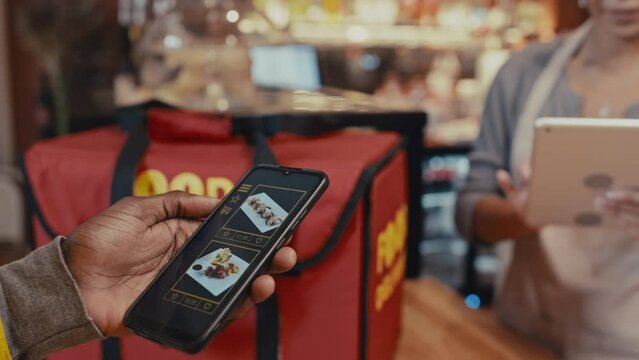 Close up shot of cellphone with delivery mobile app in hand of courier describing order to restaurant worker