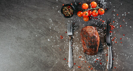 A generous piece of grilled steak on a table with fork and knife cutlery and green and red hot...