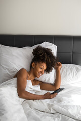 Portrait of a beautiful happy young black woman using smartphone in a comfortable bed in the morning. Afro hairstyle natural beauty holding her phone. Relaxing at home.
