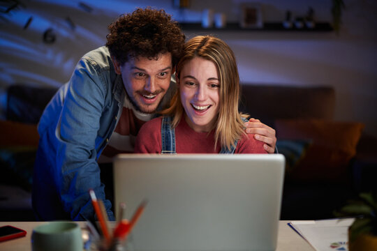 Surprised Caucasian Couple Looking Happy At Computer Sitting At Desk Living Room At Night. Excited Millennial People Having Fun At Home Using Laptop. Positive Relationships And Leisure Moments.