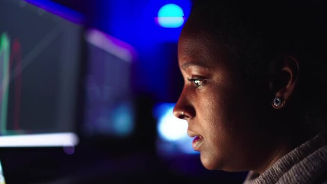 Close up serious young black woman face using laptop in darkness. Concentrated African female working online in home office with neon lights. Business, telecommuting, programming and freelancing.