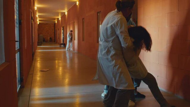 Two male doctors working in asylum restraining aggressive patient in straitjacket, taking him away to calm him down