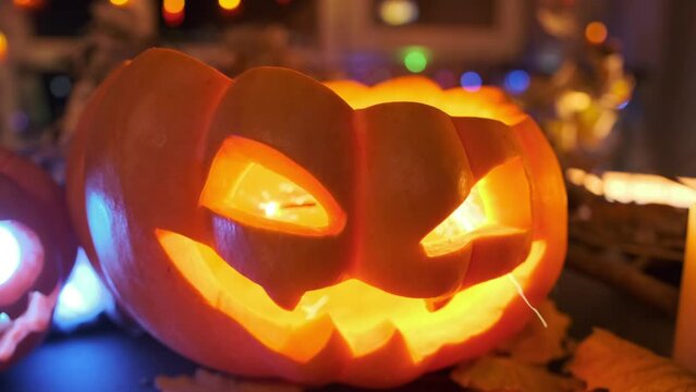 Close-up of a carved carnival pumpkin with glowing eyes being set on fire and covered with a background of carnival lights. Jack O'Lantern as a bat during Halloween celebrations