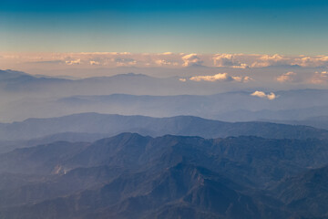 Aerial view of dusky layers of blue and green mountain ranges amid clear blue sky captured from Airplane window