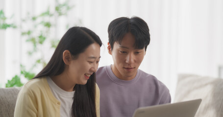 Obraz premium Portrait of a Young Korean Couple, Laptop Computer in Hand, Connecting with Friends on Social Media. Researching Online and Shopping for a Surprise Gift to Family. Sitting on a Cozy Couch in a Flat