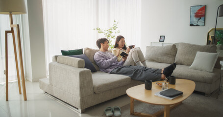 South Korean Couple Sitting on a Cozy Couch, Playing Funny Arcade Video Games Together in a Stylish Bright Living Room. Beautiful Couple in a Relationship Enjoying Time on a Sofa at Home - obrazy, fototapety, plakaty