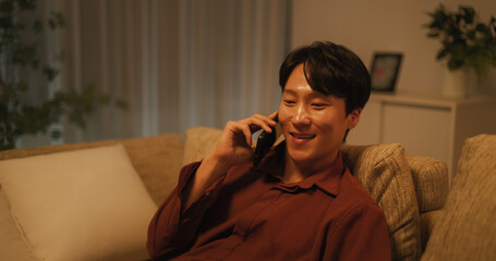 Young South Korean Man is Relieved to Get Positive News on a Phone. Happy Asian Male Smiles While Having a Conversation on a Call with a Friend. Handsome Man Resting on Couch at Home Before Sleep