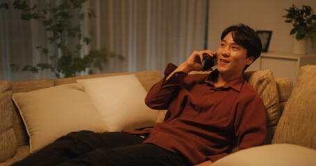 Obraz premium Handsome South Korean Young Man is Having a Joyful Phone Conversation, Having a Casual Chat with a Friend, Telling Fun Stories While Relaxing on Comfortable Sofa in the Evening