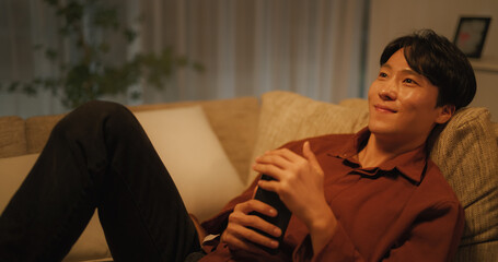 Fototapeta premium South Korean Young Man Enjoying His Leisure Time while Relaxing and Using a Smartphone. Asian Male Smiling with a Dreamy Look While Using Social Media to Talk to his Girlfriend