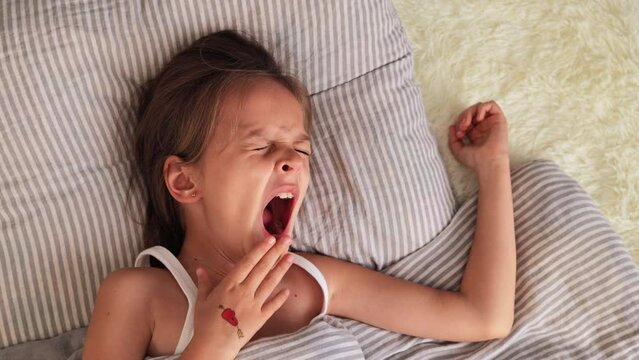 Dreaming in a comfy bed. Comfort and calm in bed. Cute girl yawning in bed at morning waking up looks sleepy covering widely open mouth lying on soft pillow needs to get up wants to have more nap.