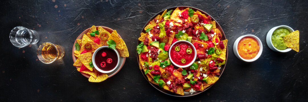 Loaded nachos panorama. Mexican nacho chips with beef, overhead flat lay shot with guacamole sauce, cheese salsa, tequila drinks, on a black slate background
