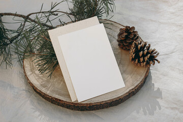 Christmas still life. Vertical greeting card, invitation mockup on cut wooden round board. Green pine tree branch in sunlight. Grey linen tablecloth. Festive winter stationery template. Flat lay, top