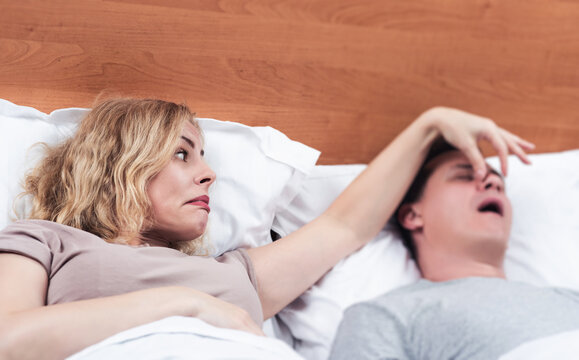 Two in bed, a woman tries to make a man stop snoring by covering his nose with her fingers. Snoring as a problem.