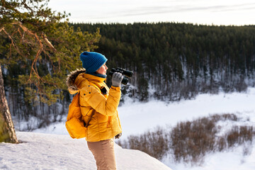 Fototapeta na wymiar Young woman in yellow looking through binoculars at birds on snowy river against winter forest Birdwatching, zoology, ecology Research in nature, observation of animals Ornithology copy space