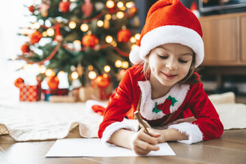 Little girl in Santa's hat lying by the New Year tree, writing her heartfelt letter to Santa Claus