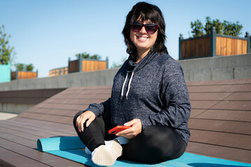 woman in sportswear and sunglasses sits on a mat in the park with a phone in her hands. Smiling at...