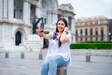 Happy young ethnic mexican woman taking selfie and top looking at screen of smartphone while sitting in front of blurred Bellas Fine Arts Palace in Mexico City and wearing traditional clothes