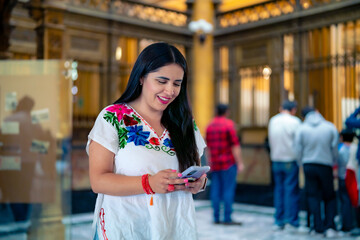 Happy young mexican woman tourist in embroidered top looking at screen of smartphone while standing in Postal palace in downtown Mexico City and browsing pictures in light against blurred interior