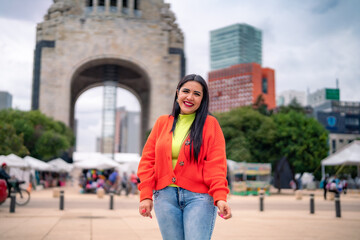 Fototapeta premium Smiling young Hispanic ethnic woman tourist smiling and looking at camera while standing in street in front of blurred Monument to the Revolution in Mexico city against cloudy blue sky in daylight