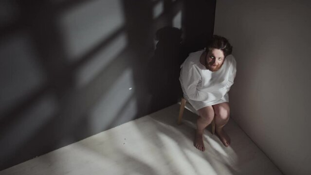 High angle shot of Caucasian man with schizophrenia wearing straitjacket sitting on chair in corner of ward grimacing, laughing and shaking head