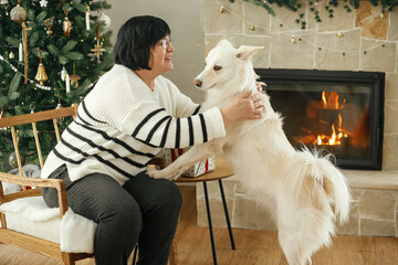 Beautiful mature woman playing with cute white dog on background of stylish christmas tree and festive fireplace. Happy adult woman enjoying cozy winter holiday time with pet