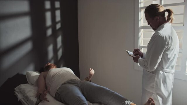 Professional psychiatrist standing in ward looking after patient with mental disorder and making notes in medical record