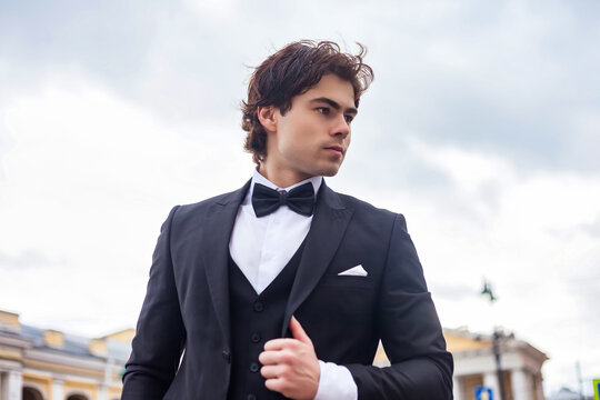 Portrait of confident stylish man in tuxedo and bow tie posing at cloudy sky, strong looking away. Pensive fashionable young guy businessman in tux, luxury image cinematic style. Copy ad text space