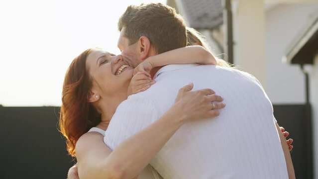 American dream family outdoors near their house. A man holds his daughter in arms and goes to wife. Father, mother and daughter hug and kiss with tenderness and warmth in their hearts. The concept of