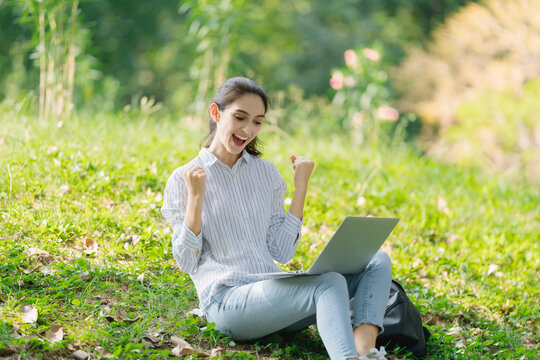 Young woman smiles and laughs celebrating joy by raising her hands with a laptop under a tree in the park. Woman working online using computer or video conferencing