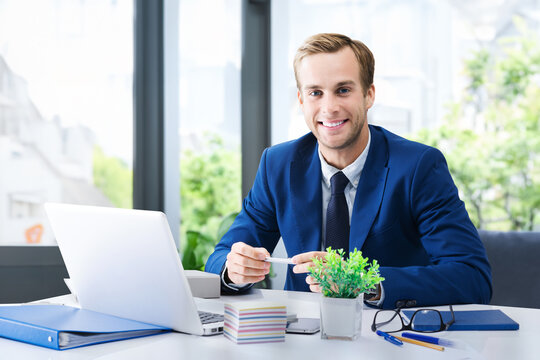 Portrait photo - business man at workplace table near window on background. Happy smile confident businessman in blue suit work with laptop computer. Business lawyer, bank consulting, trader
