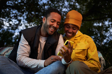 Happy young African American couple using mobile outdoors. Two joyful friends sitting looking at yellow phone smiling at dusk in winter. Online relationships generation z nature getaways on vacation.