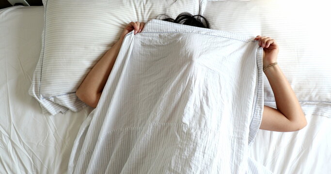 Fun young woman playing peekaboo with bed blanket. Girl gide and seek in the morning