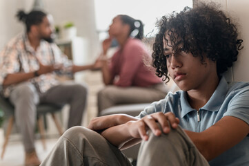 A teenage boy is sitting on the floor looking sad while his parents are arguing and fight - obrazy, fototapety, plakaty