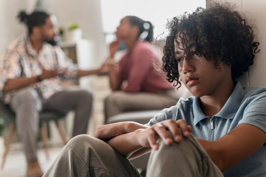 A teenage boy is sitting on the floor looking sad while his parents are arguing and fight