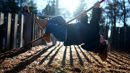 One playful child hangs upside down at playground in park during sunny autumn day. Child playing outdoors with sunlight flare