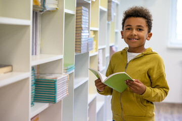 Cute dark-skinned boy in hoodie with a book at school premises