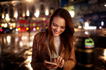 Happy young caucasian woman using a phone in the city at night