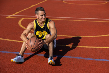 Smiling attractive man sitting on the basketball court after playing game, resting after match.
