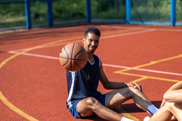 Cheerful man resting sitting on the basketball court after playing game, holding out ball.