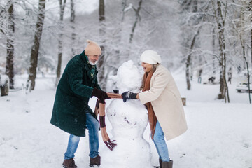 Elegant senior couple building snowman during cold winter snowy day.