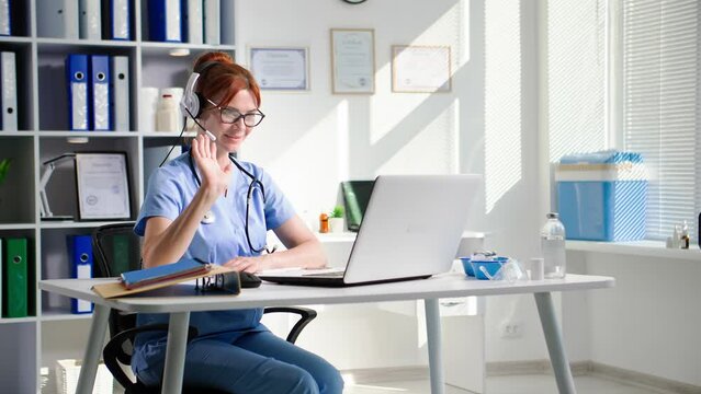 consultation with doctor, young female medical worker in glasses talks on video with a patient using a headset while sitting at a table with a laptop, smiling and looking at camera
