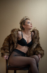 Attractive blonde model with pantyhose, black bra and fur coat posing provocatively on chair. Fashion portrait of sensual blonde, studio shot. Sensual female in black lingerie posing against wall.