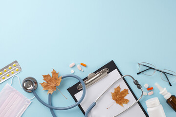 Take medications prescribed by your doctor in autumn. Top view shot of clipboard, stethoscope, thermometer, medical mask, eyeglasses, pills, fallen leaves on soft blue background with ad area