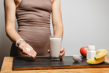 Athletic woman in sportswear holding glass of protein drink cocktail and white capsules of amino acids on a table with measuring spoon of protein powder and fruits